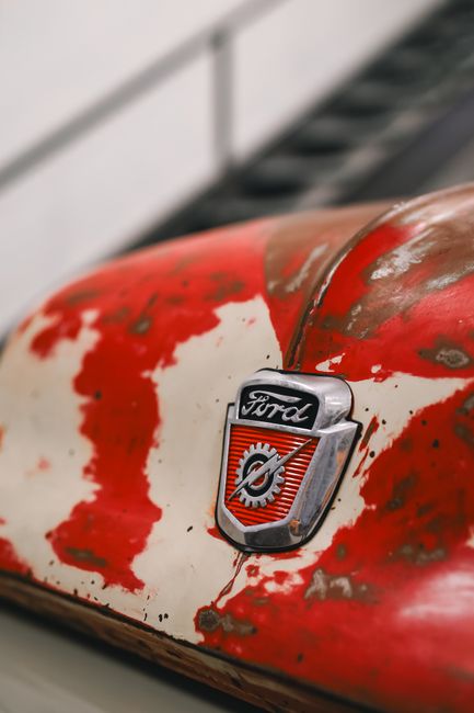 Close-up image of a vintage Ford vehicle with a weathered, chipped red and white paint finish, displaying a central Ford emblem attached to the hood. The emblem features a silver border with the Ford 