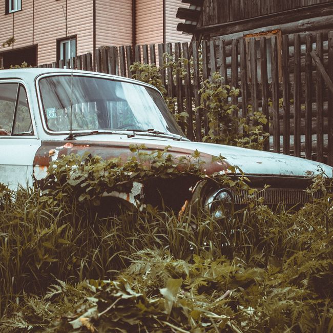 Close-up image of a vintage Ford vehicle with a weathered, chipped red and white paint finish, displaying a central Ford emblem attached to the hood. The emblem features a silver border with the Ford 