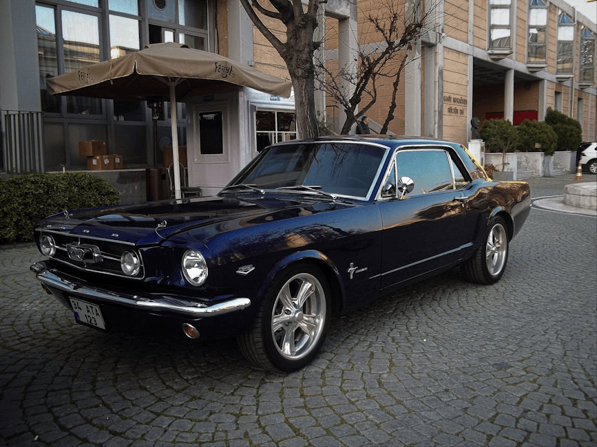 A classic dark blue Ford Mustang Fastback parked on a cobbled street in an urban setting, with a building featuring large glass windows and a sidewalk with trimmed hedges and trees in the background. 